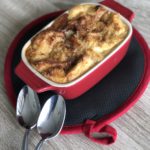 Mini red dutch oven filled with bread pudding sitting on a red and black hot plate with two spoons against a grey wood-patterned background